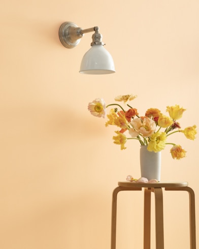 A Delicate Peach-painted wall with single sconce, and a wooden stool with flower-filled vase.